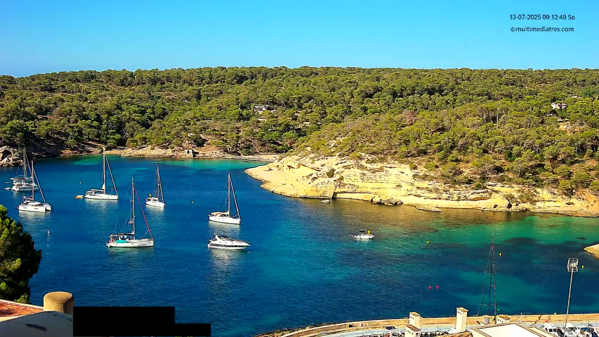 Several sailboats are anchored in a calm, clear turquoise cove surrounded by a lush, green, hilly coastline under a bright sunny sky.