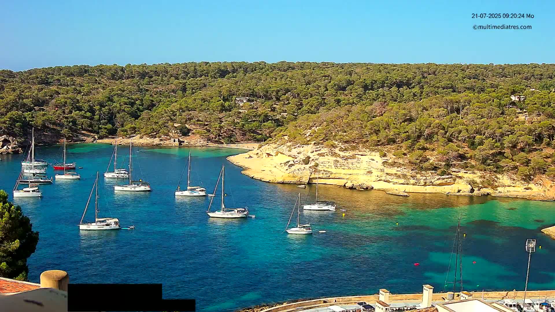 Numerous sailboats are anchored in a calm, clear turquoise bay surrounded by a lush green, hilly coastline under a bright, sunny sky.