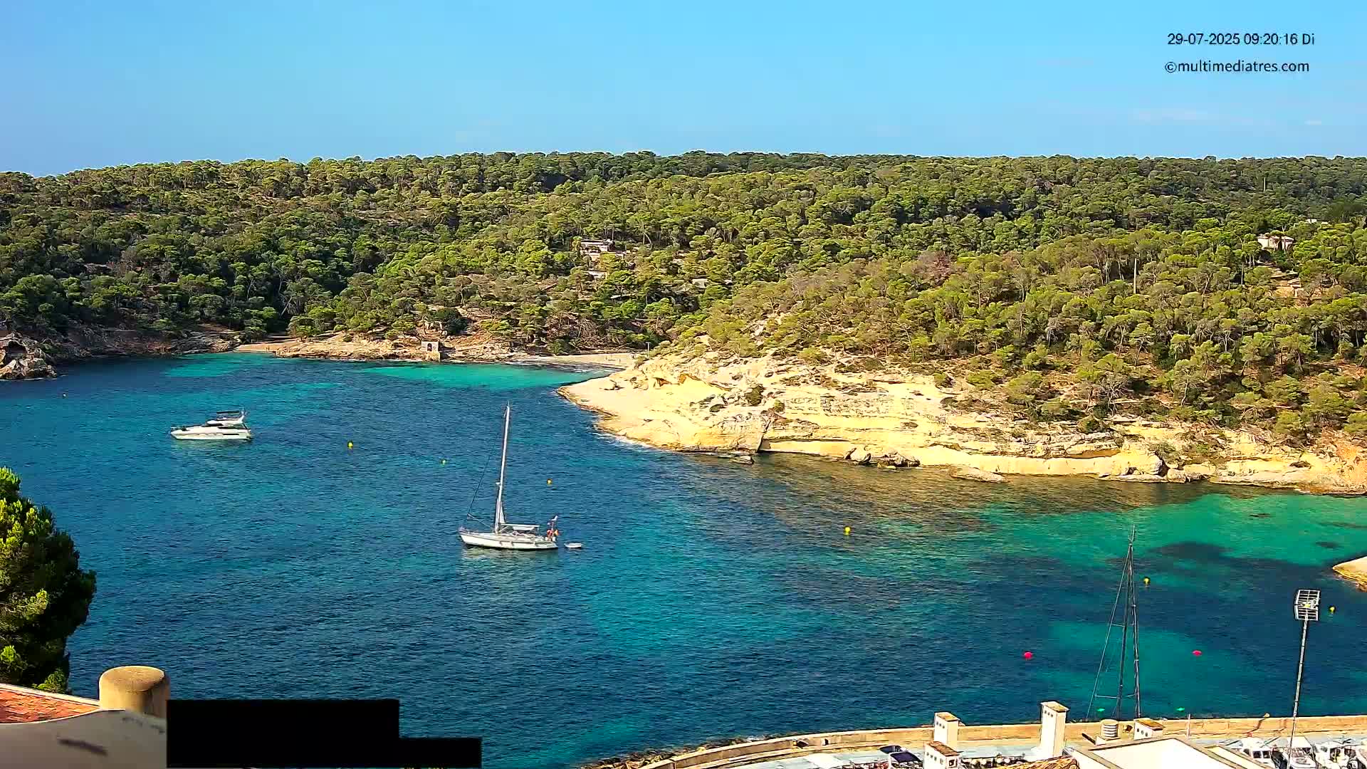 A secluded cove with turquoise water, surrounded by lush green vegetation under a clear blue sky, features several sailboats and motorboats.