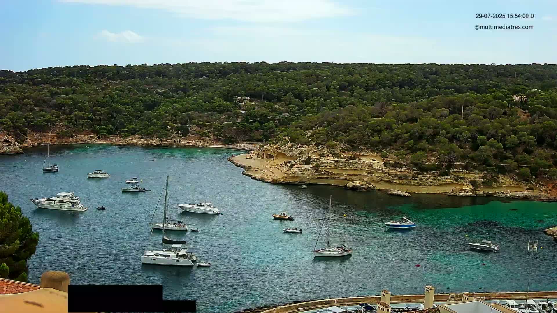 A secluded cove with turquoise water, surrounded by lush green vegetation under a clear blue sky, features several sailboats and motorboats.