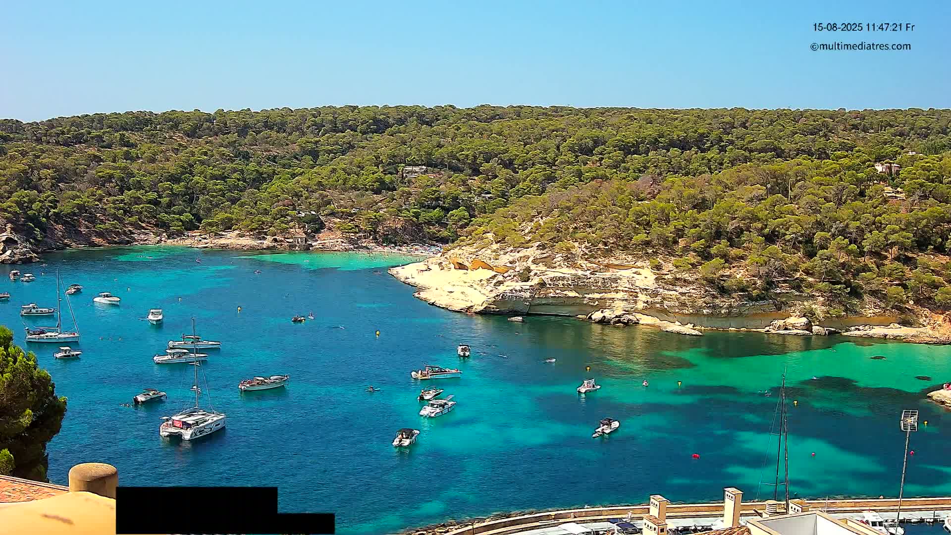 A secluded cove with turquoise water, surrounded by lush green vegetation under a clear blue sky, features several sailboats and motorboats.
