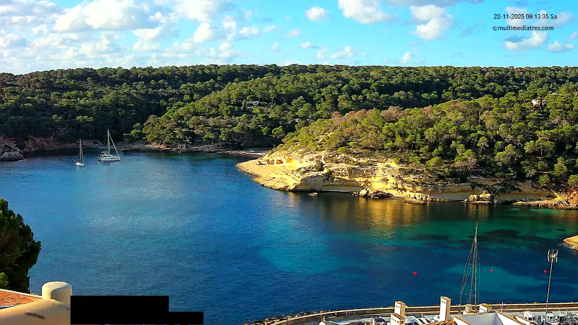 A tranquil bay with clear blue-turquoise water is nestled between lush green, pine-covered hills and sandy-rocky cliffs, featuring a few anchored sailboats and a partial marina in the foreground, all under a bright, partly cloudy sky.