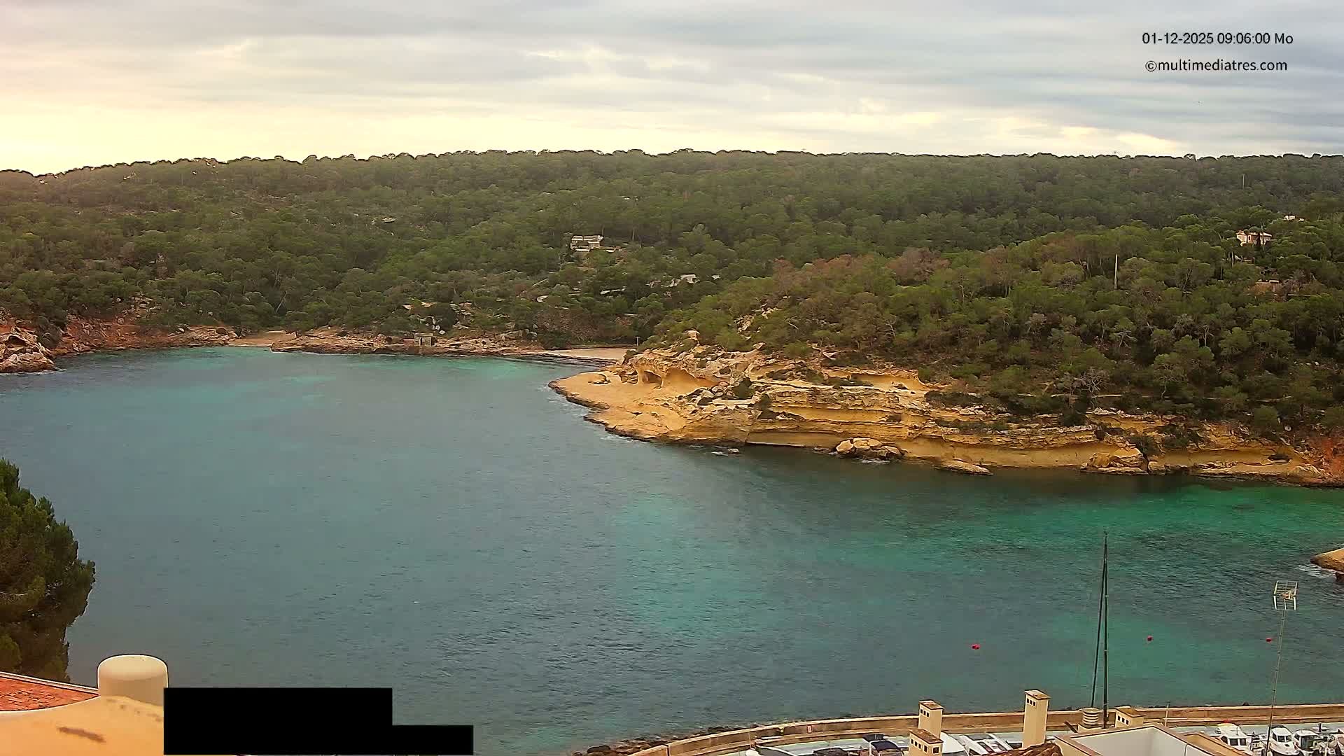 A tranquil coastal bay with clear turquoise waters is bordered by lush green hills and golden-brown rocky cliffs, viewed under an overcast sky, with rooftops and docked boats partially visible in the foreground.