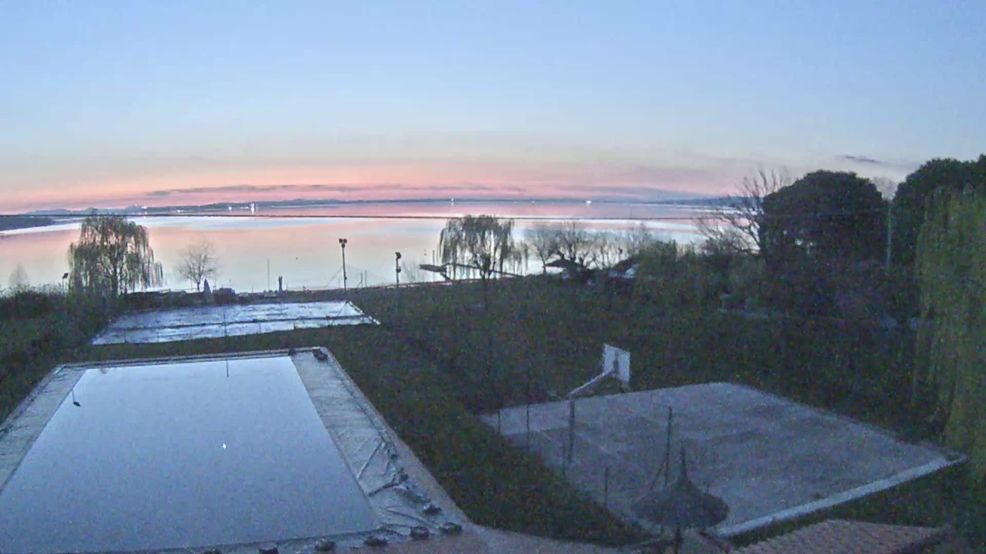 A calm lake at dawn is visible from a property containing an empty swimming pool and a basketball court.