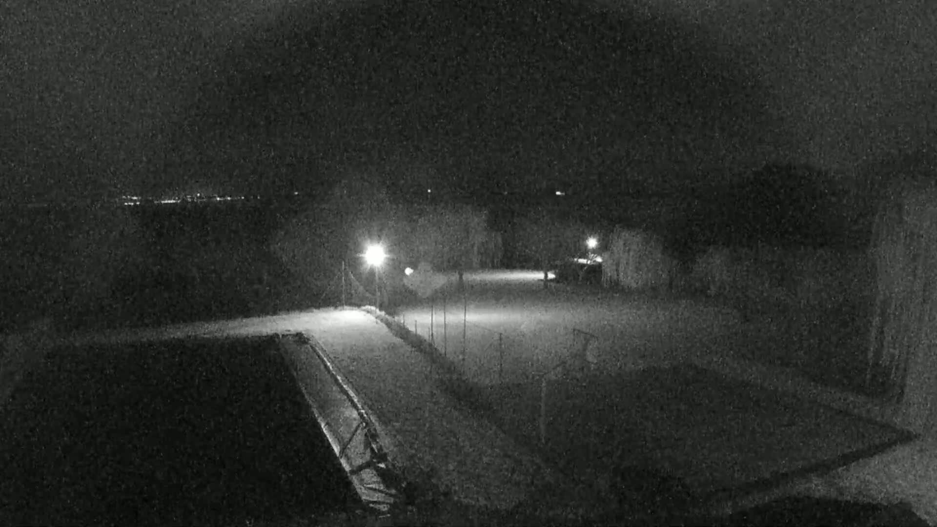 A nighttime, snow-covered outdoor scene shows a lit area with what appears to be a sports field and a dark pool or similar structure, with distant lights visible in the background.