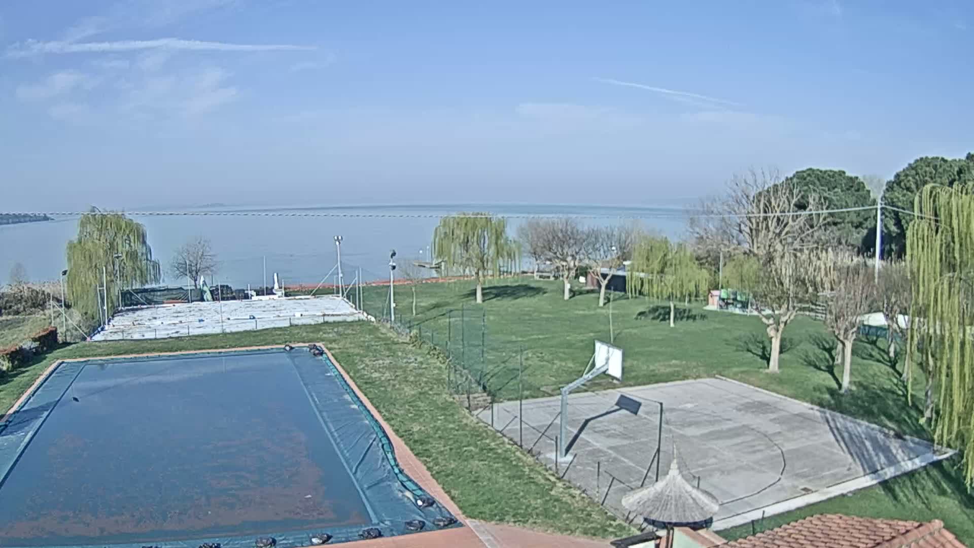 A covered swimming pool and an outdoor basketball court sit on a grassy area overlooking a calm lake under a clear, sunny sky.