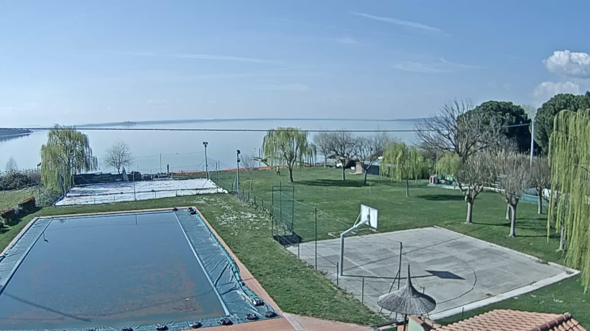 An outdoor scene on a sunny day shows a covered swimming pool, a basketball court, and a grassy area overlooking a calm lake.