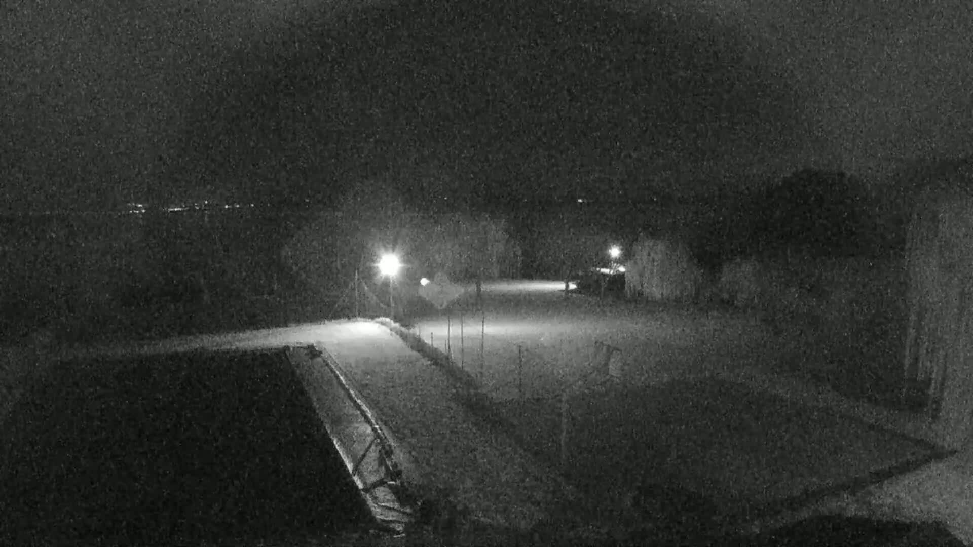 A nighttime, snow-covered outdoor area is illuminated by several lights, showing a paved path and fenced-in sections.