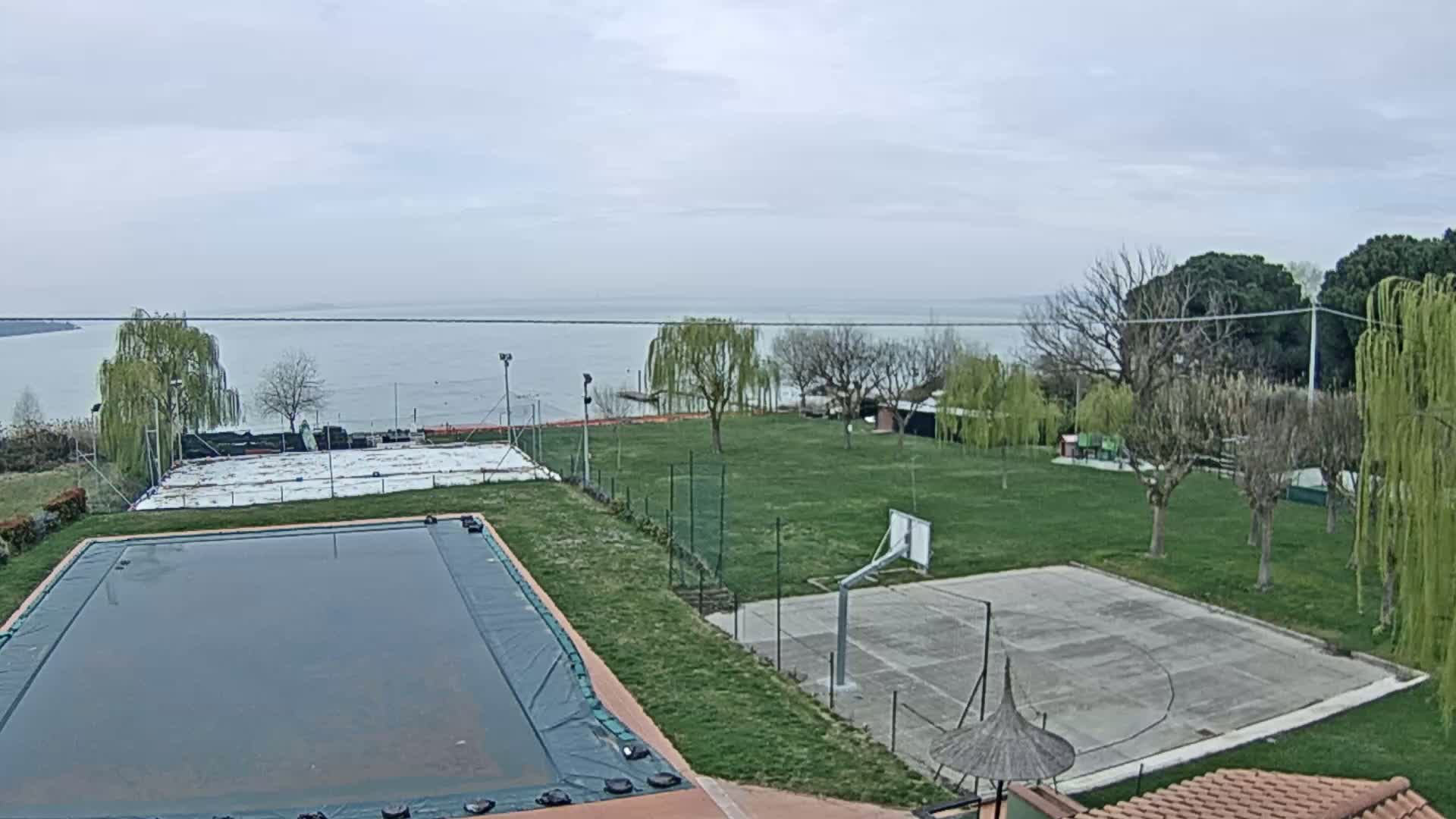 An outdoor view shows a covered swimming pool, a basketball court, and a tennis court on a grassy area overlooking a calm lake under a cloudy sky.