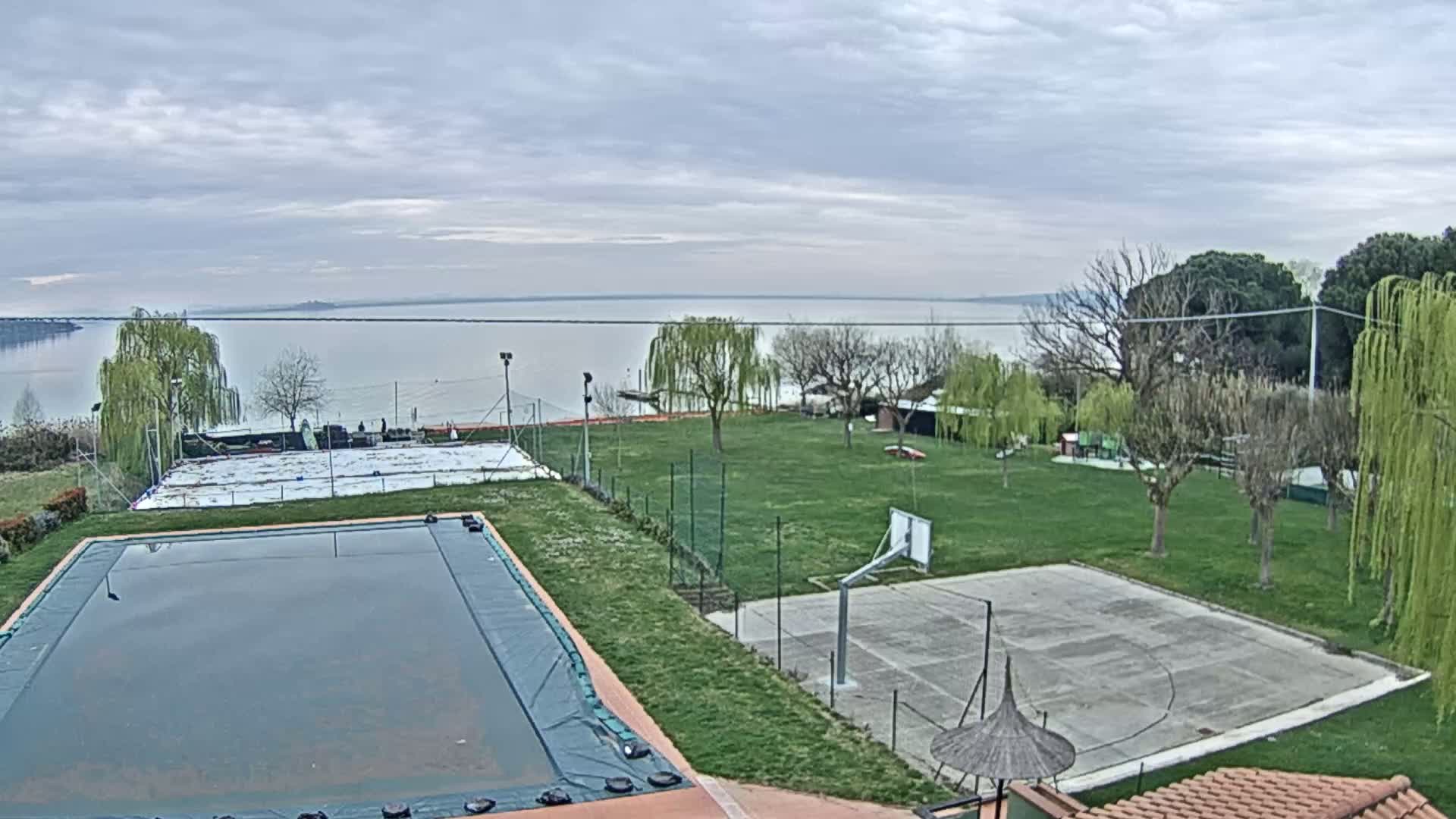 A covered swimming pool and a basketball court are situated on a grassy area overlooking a calm lake under a cloudy sky.