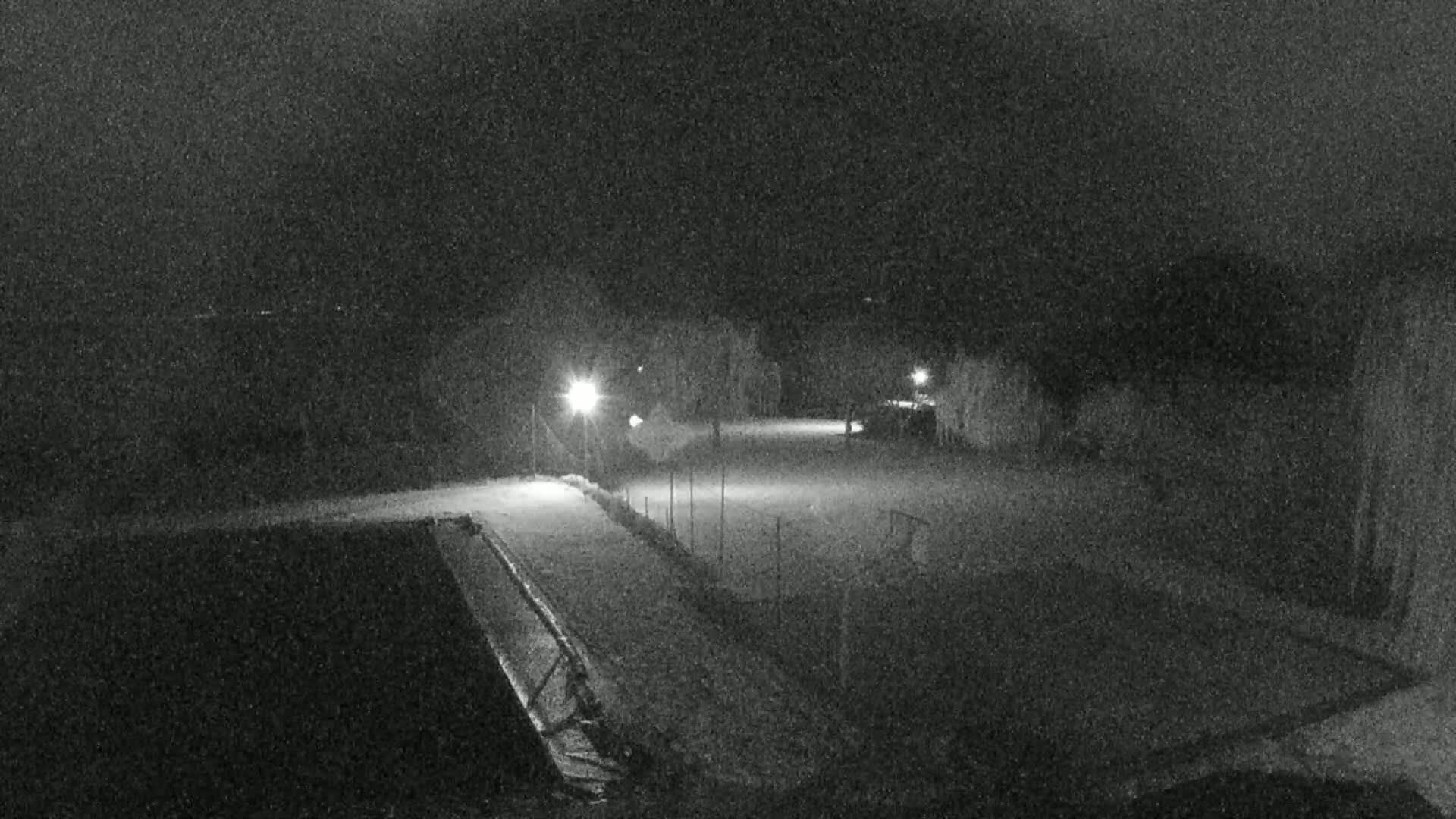 A nighttime, snow-covered parking lot is illuminated by several lights, with a fenced-off area visible to the right.