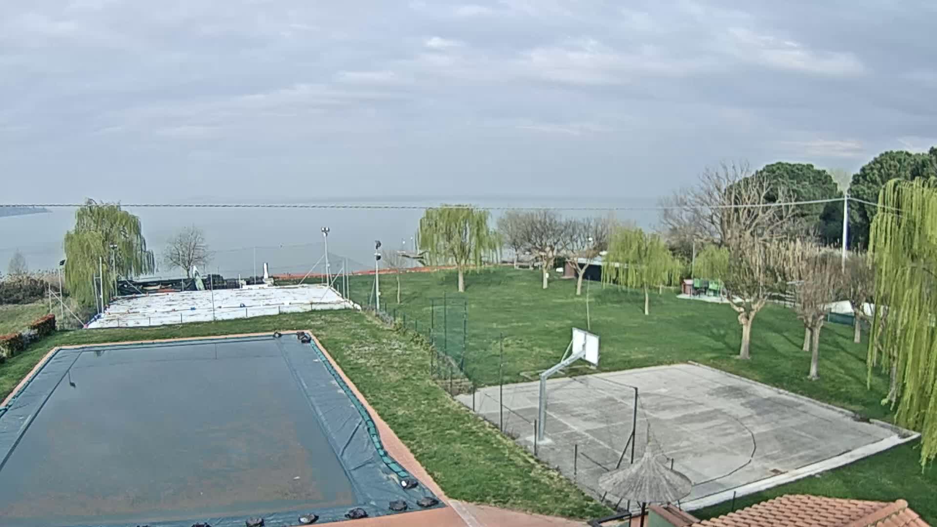 An outdoor view of a covered pool, a basketball court, and a grassy area with trees, overlooking a calm body of water under a cloudy sky.