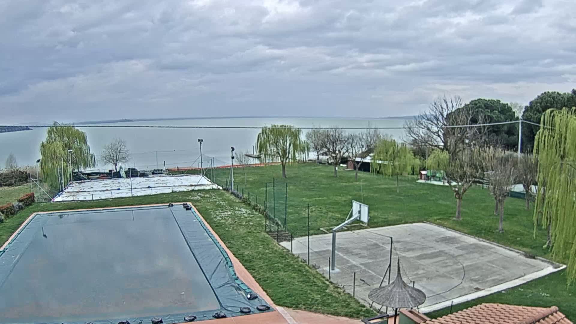 A covered swimming pool and a basketball court sit on a grassy area overlooking a calm lake under a cloudy sky.