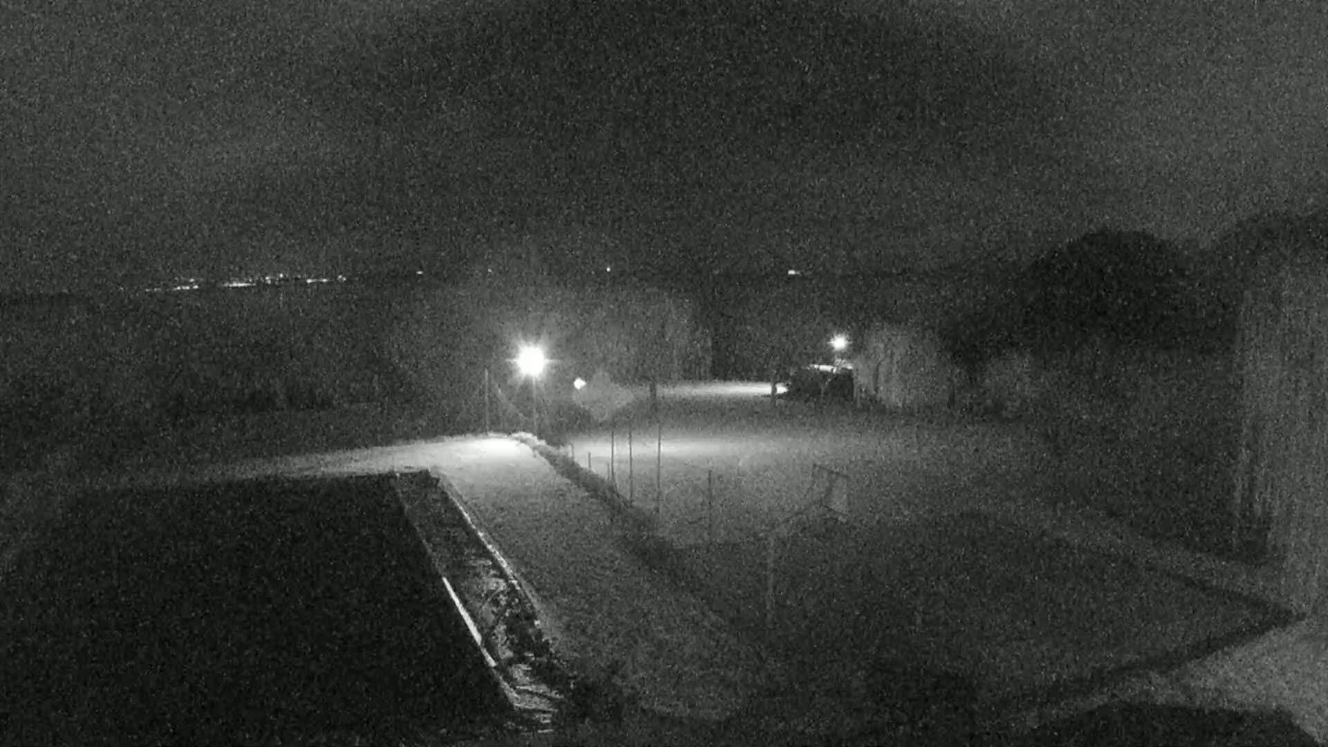 A snow-covered nighttime scene shows an empty, fenced-in area illuminated by a few lights, with a building and a dark, possibly water-filled, rectangular structure in the foreground.