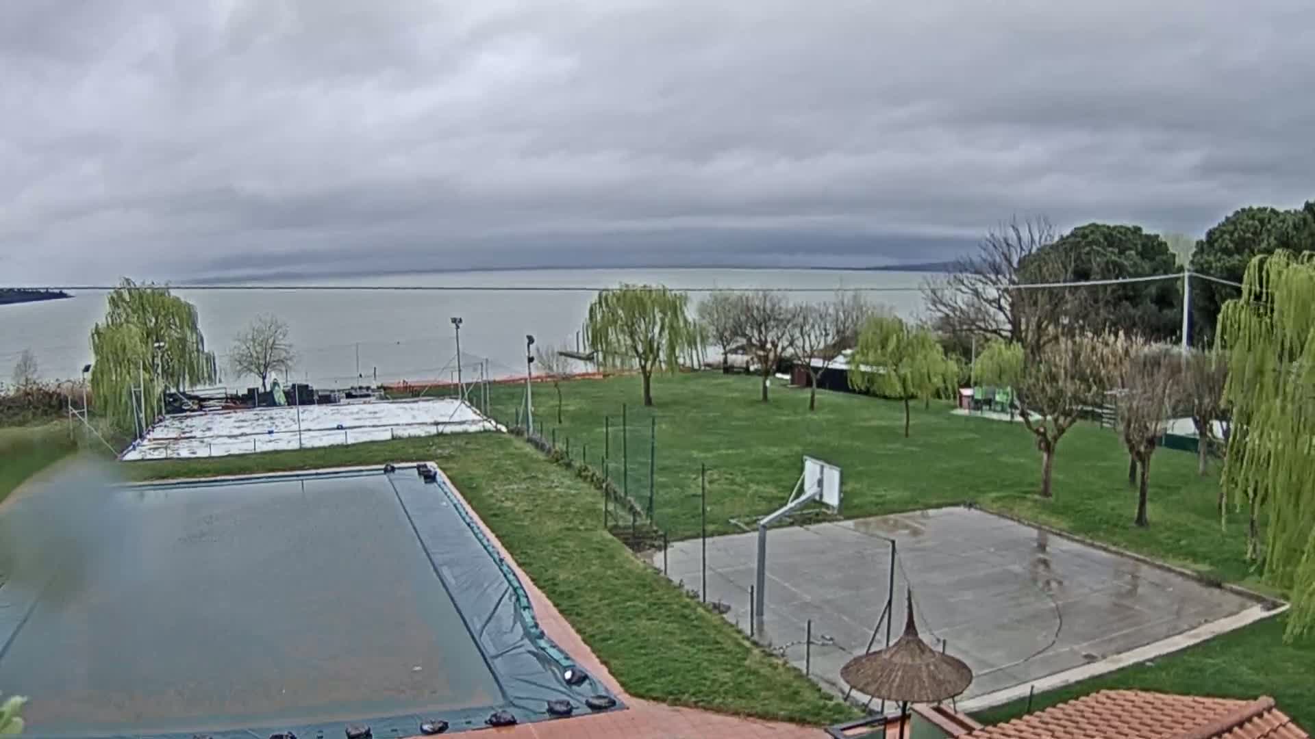 An outdoor scene shows a covered swimming pool and a basketball court on a grassy area near a large body of water under an overcast sky.