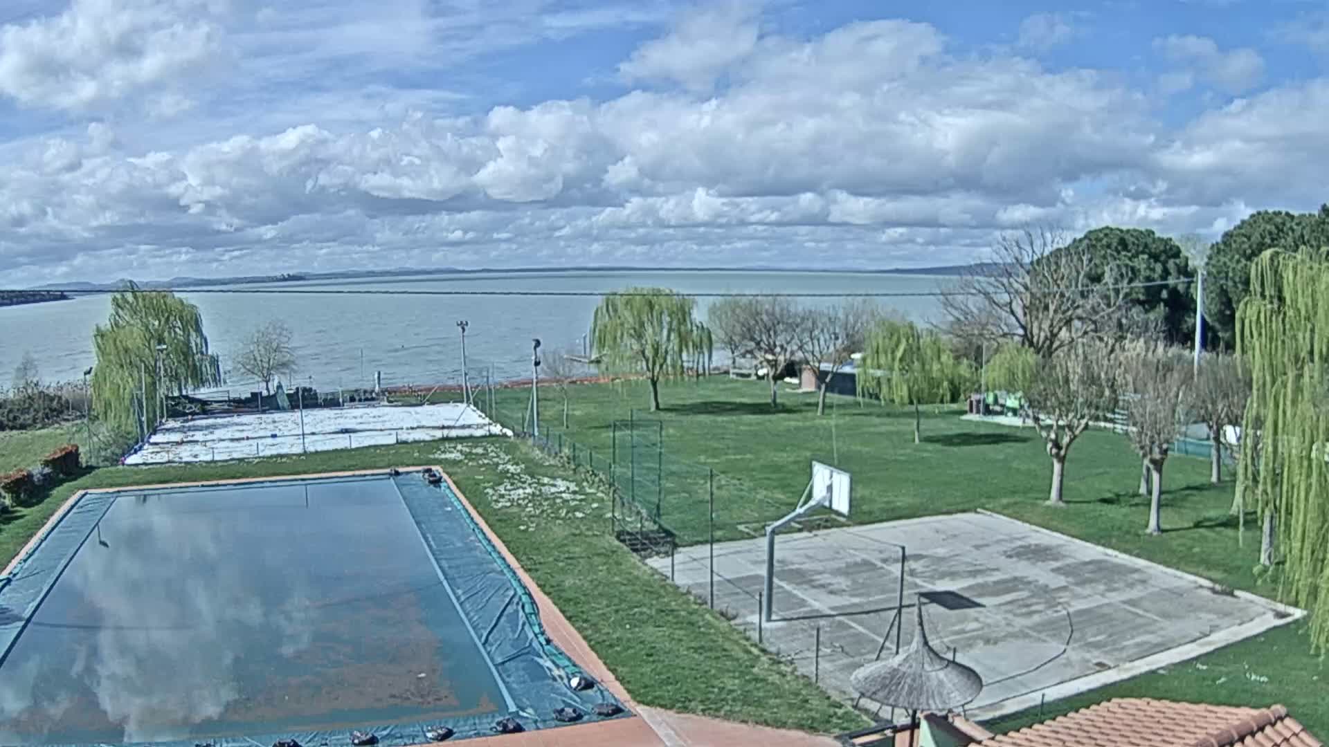A swimming pool and basketball court are situated on a grassy area overlooking a calm lake under a partly cloudy sky.