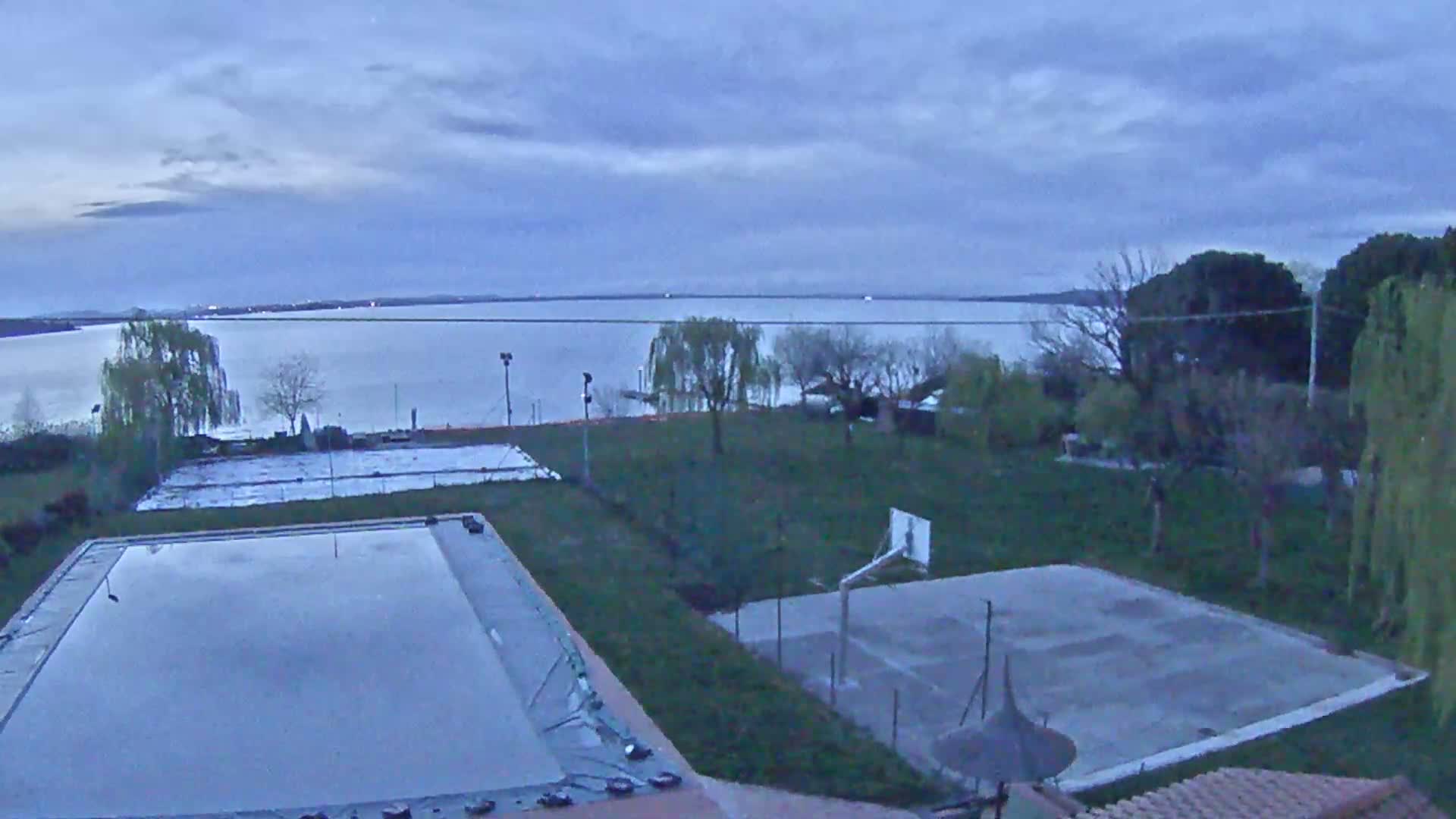 An outdoor view of an empty swimming pool and basketball court on a grassy area overlooking a calm lake under a cloudy, twilight sky.