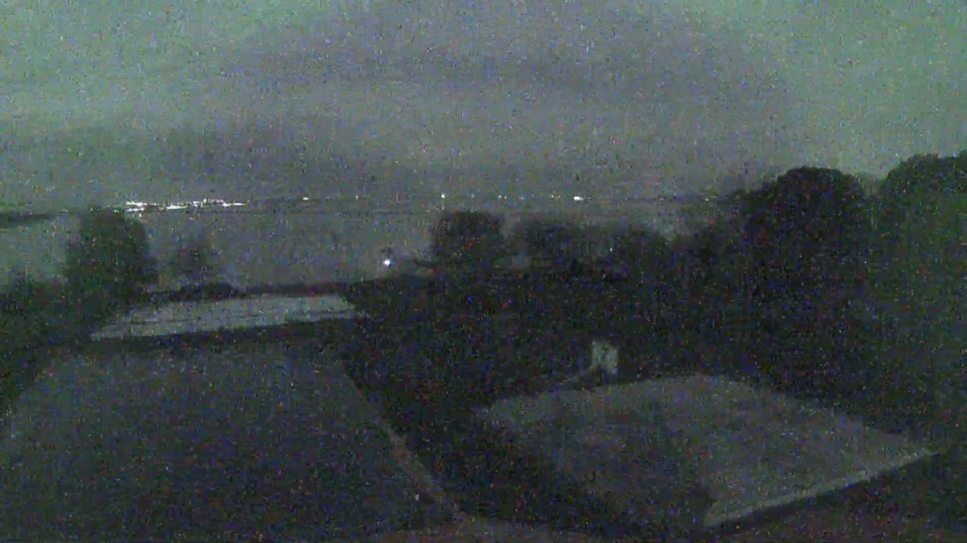A nighttime, overcast view shows dark rooftops and trees, with a body of water and distant lights visible in the background.