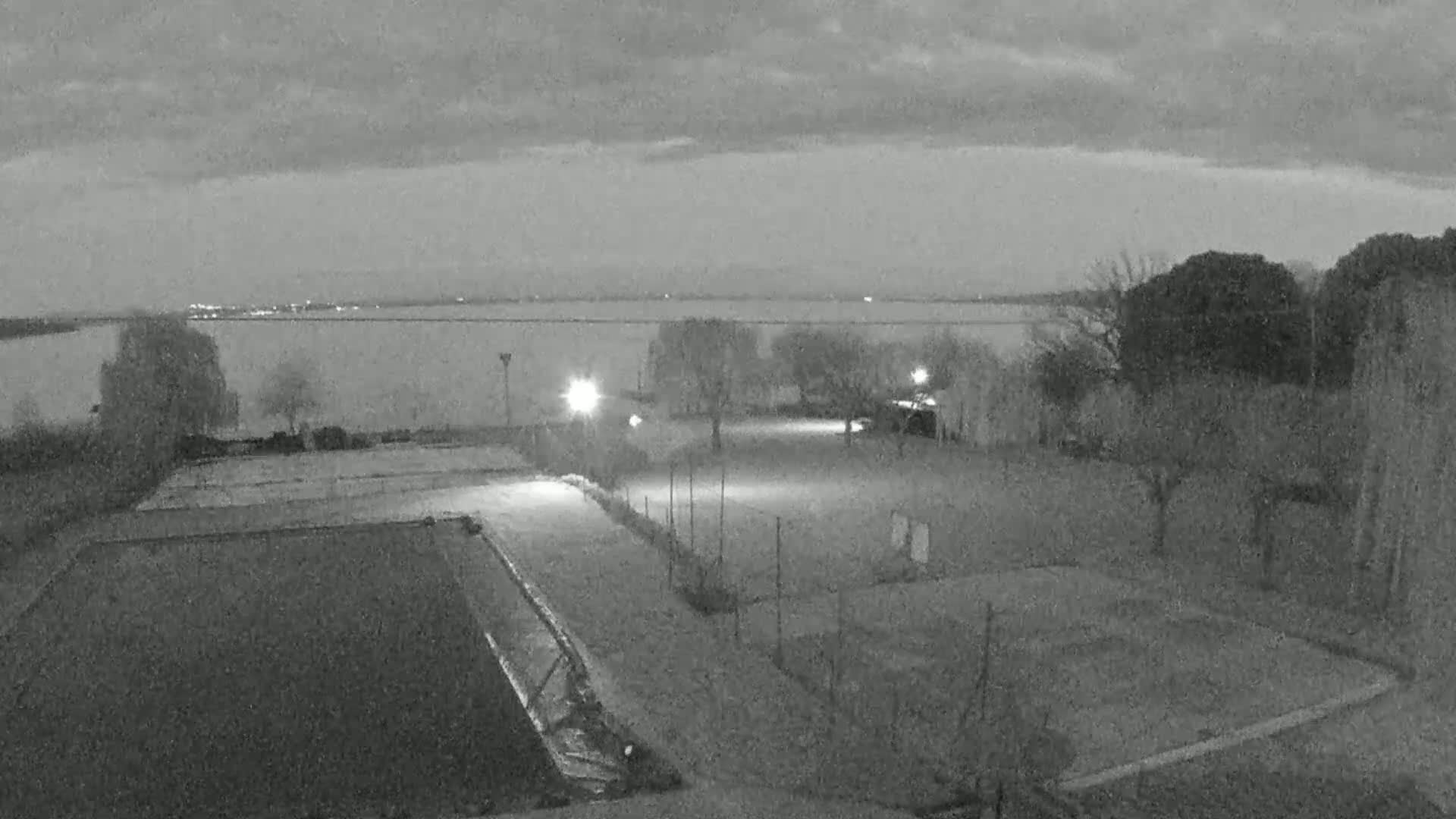 A nighttime, overcast view of empty outdoor sports courts near a body of water with sparsely spaced trees and illuminated areas.