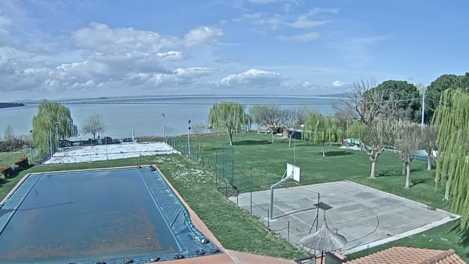 A covered swimming pool and basketball court sit on a grassy area overlooking a calm lake under a partly cloudy sky.