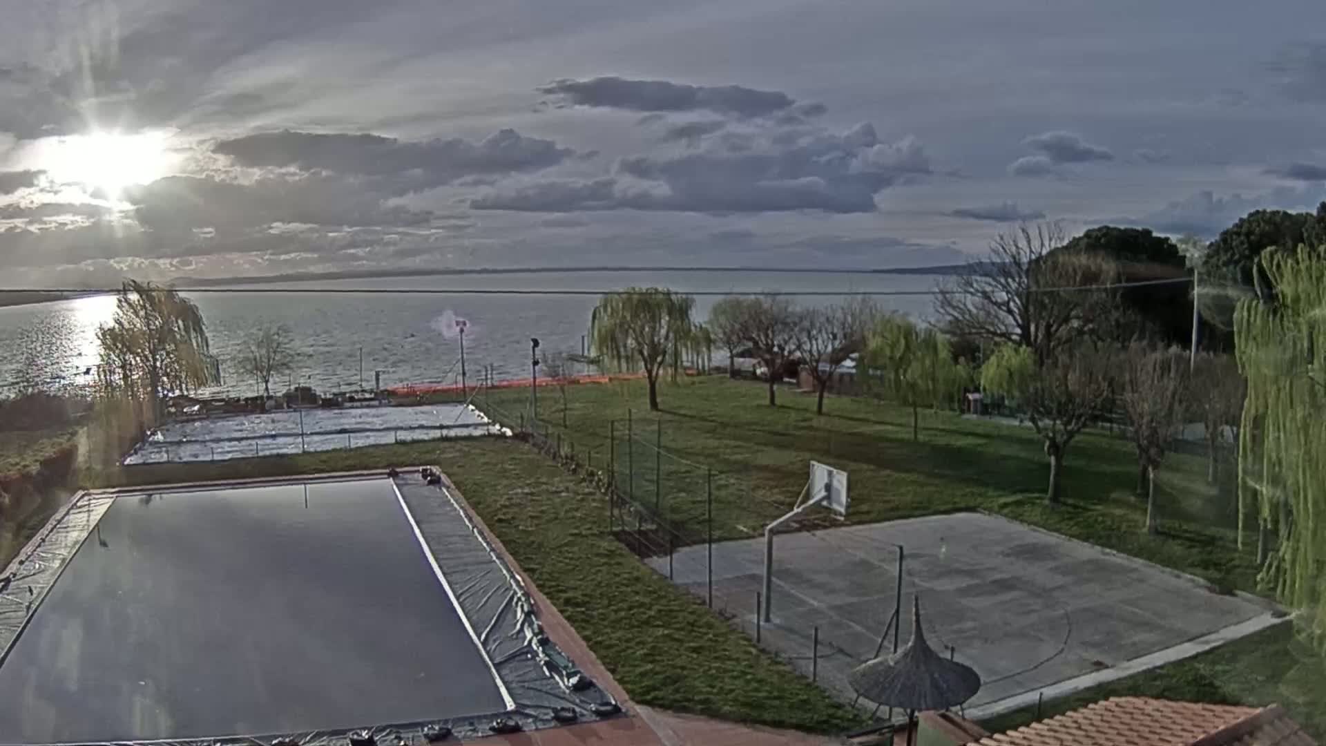 A partly cloudy sunset over a lake is visible in the background, overlooking an outdoor area with a covered swimming pool, a basketball court, and several trees.
