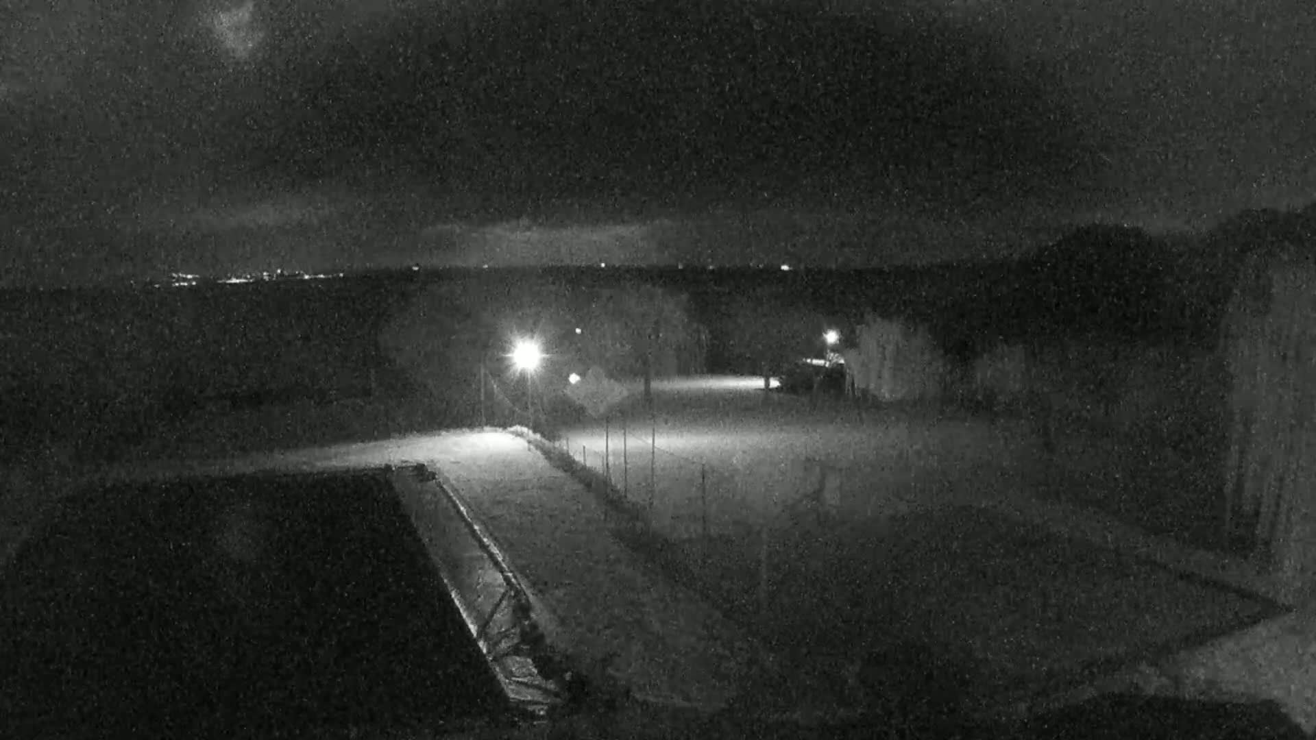 A nighttime, snowy scene shows a dark, empty paved area illuminated by several lights, a dark pool of water to the left, and a distant city skyline faintly visible on the horizon.