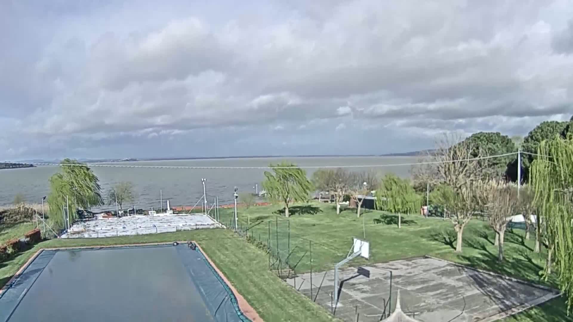 A swimming pool and basketball court are situated on a grassy area overlooking a calm lake under a partly cloudy sky.