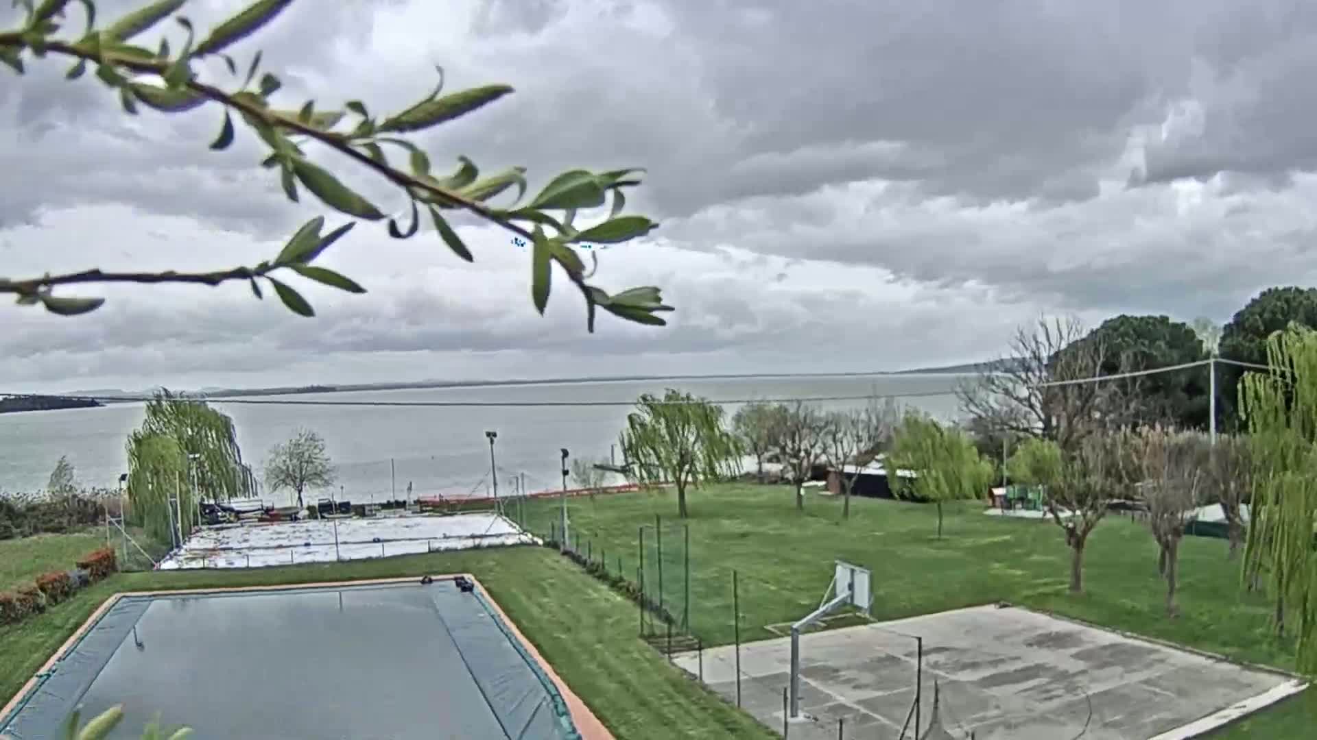 A covered pool and a basketball court are situated on a grassy area overlooking a calm lake under a cloudy sky.