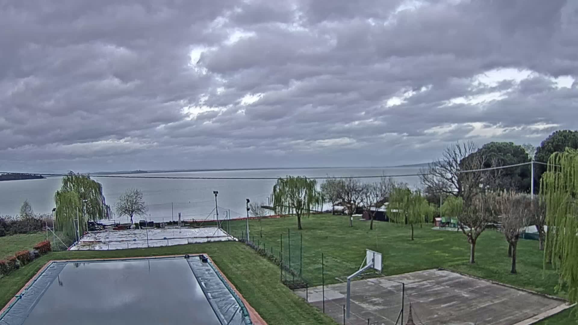 A mostly cloudy sky overlooks a calm lake,  with a grassy area containing a swimming pool and basketball court in the foreground.