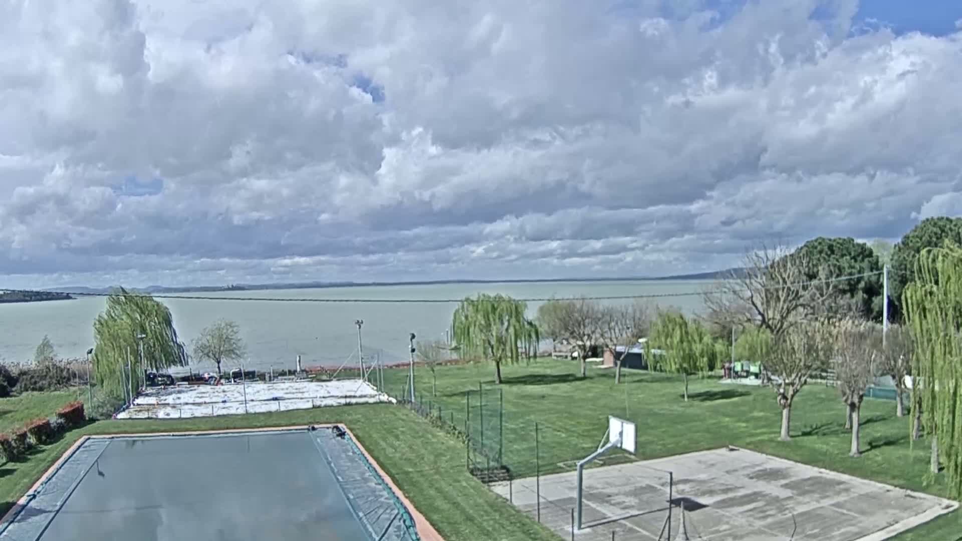 A covered swimming pool and a basketball court sit on a grassy area overlooking a calm lake under a partly cloudy sky.