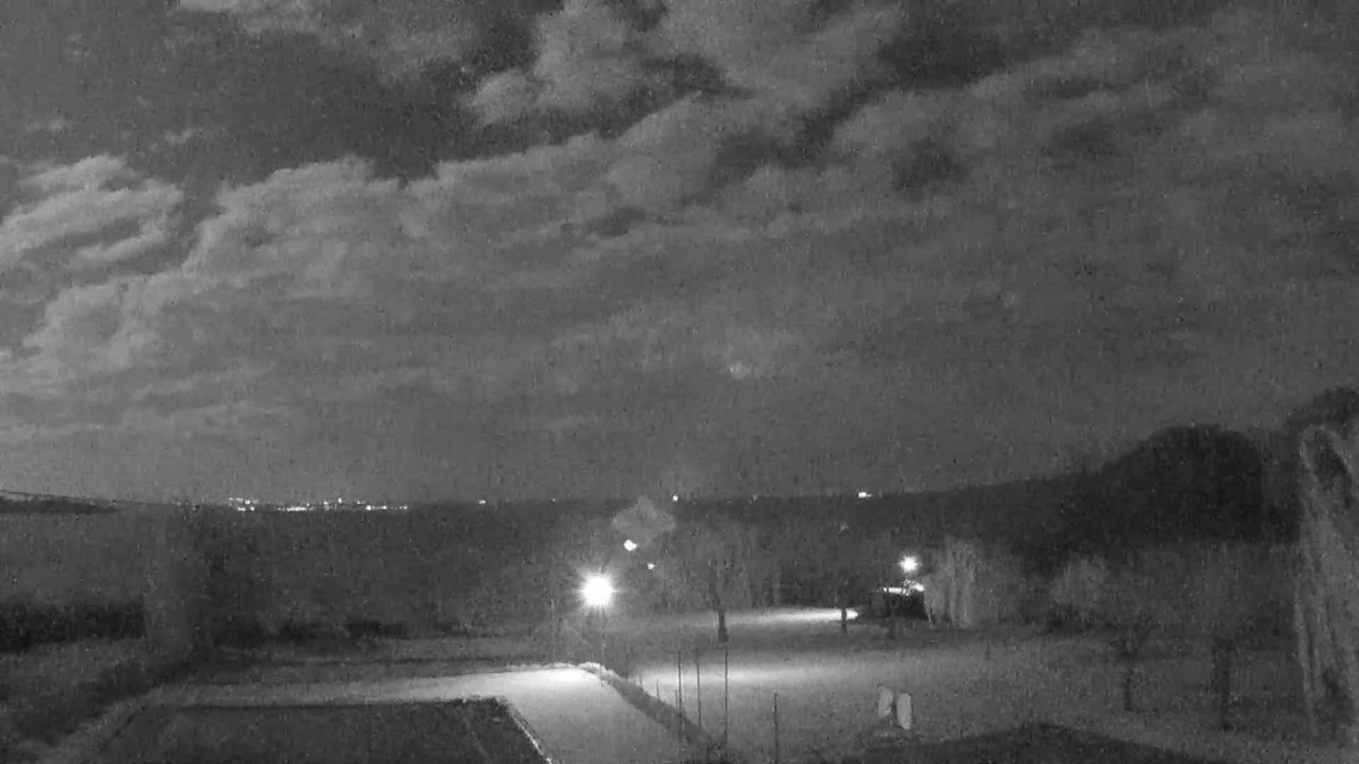 Under a partly cloudy night sky, illuminated lights shine on a paved area with sparse trees and a distant cityscape.