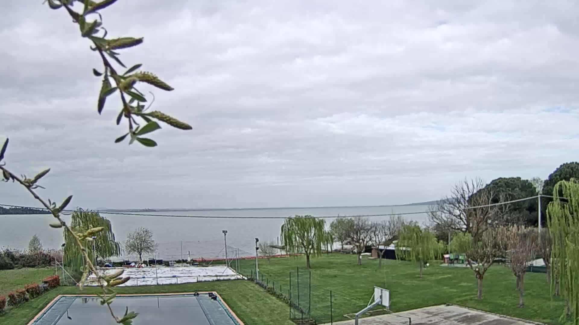 A mostly cloudy day overlooks a calm lake, with a grassy area containing a basketball court and swimming pool in the foreground.