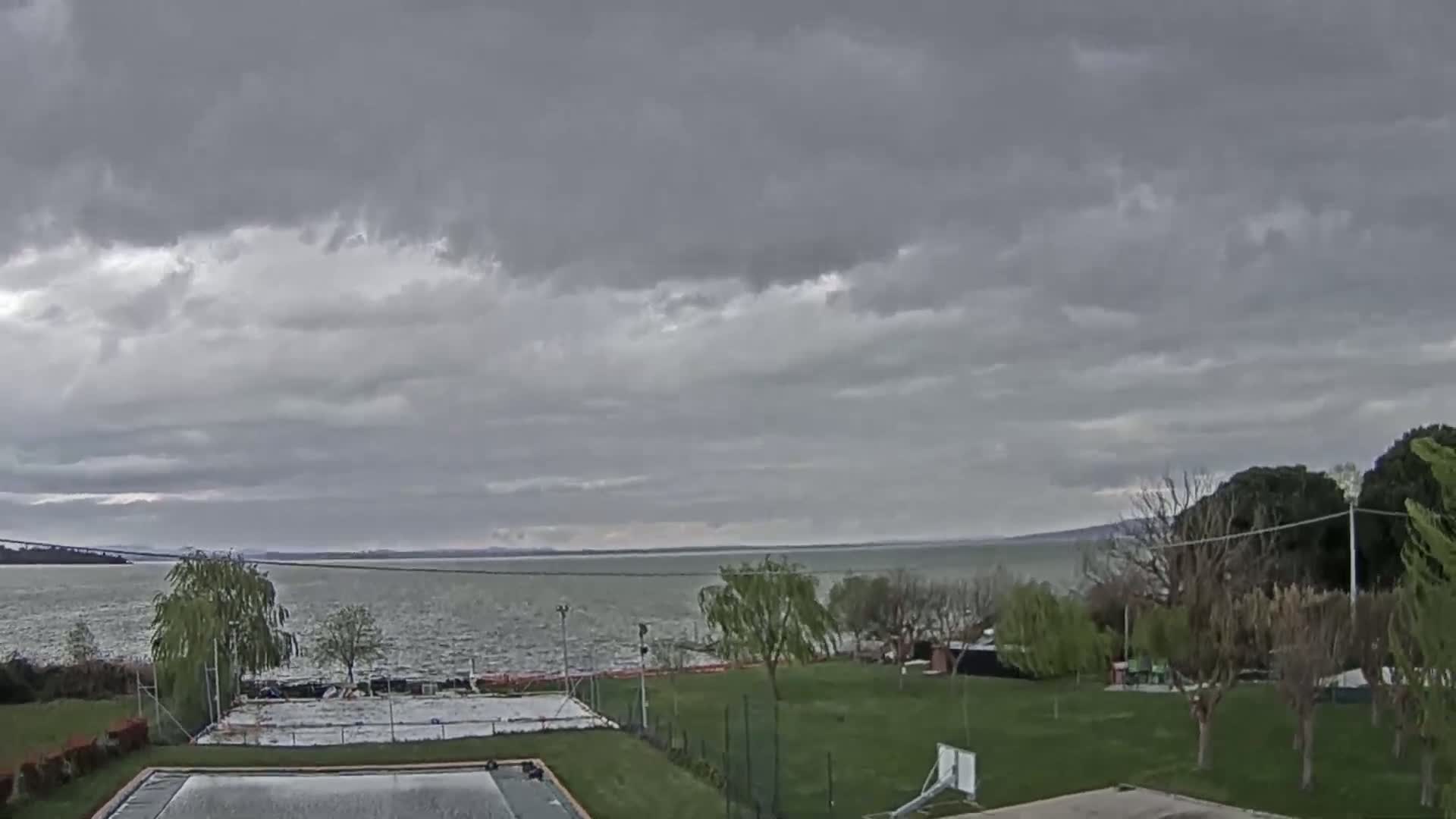 A mostly cloudy day overlooks a calm lake and grassy area with a basketball court and pool.
