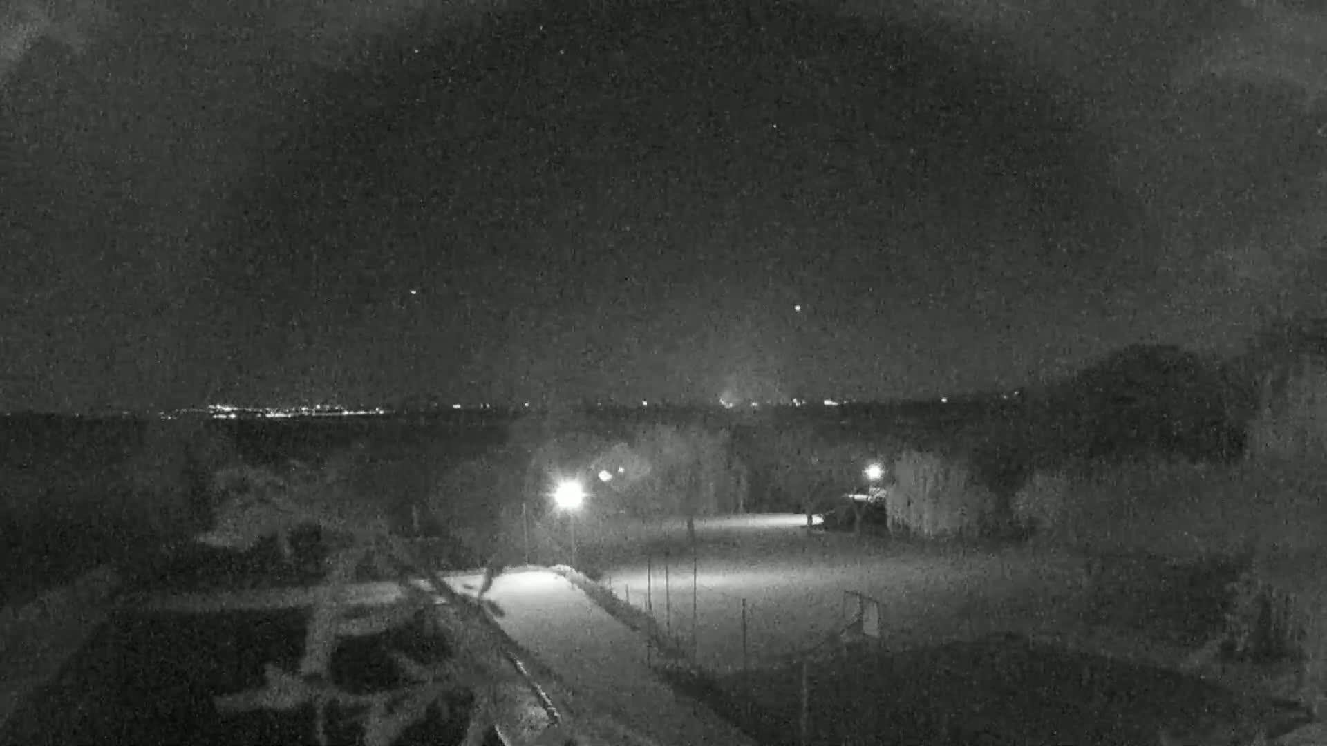 A nighttime, snow-covered outdoor scene shows a paved area lit by lamps, with trees and a distant city skyline visible under a starry sky.
