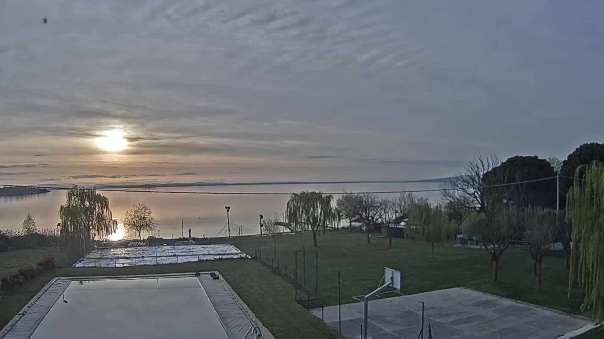 A calm lake at sunrise is visible from a vantage point overlooking a grassy area with a covered pool and basketball court.