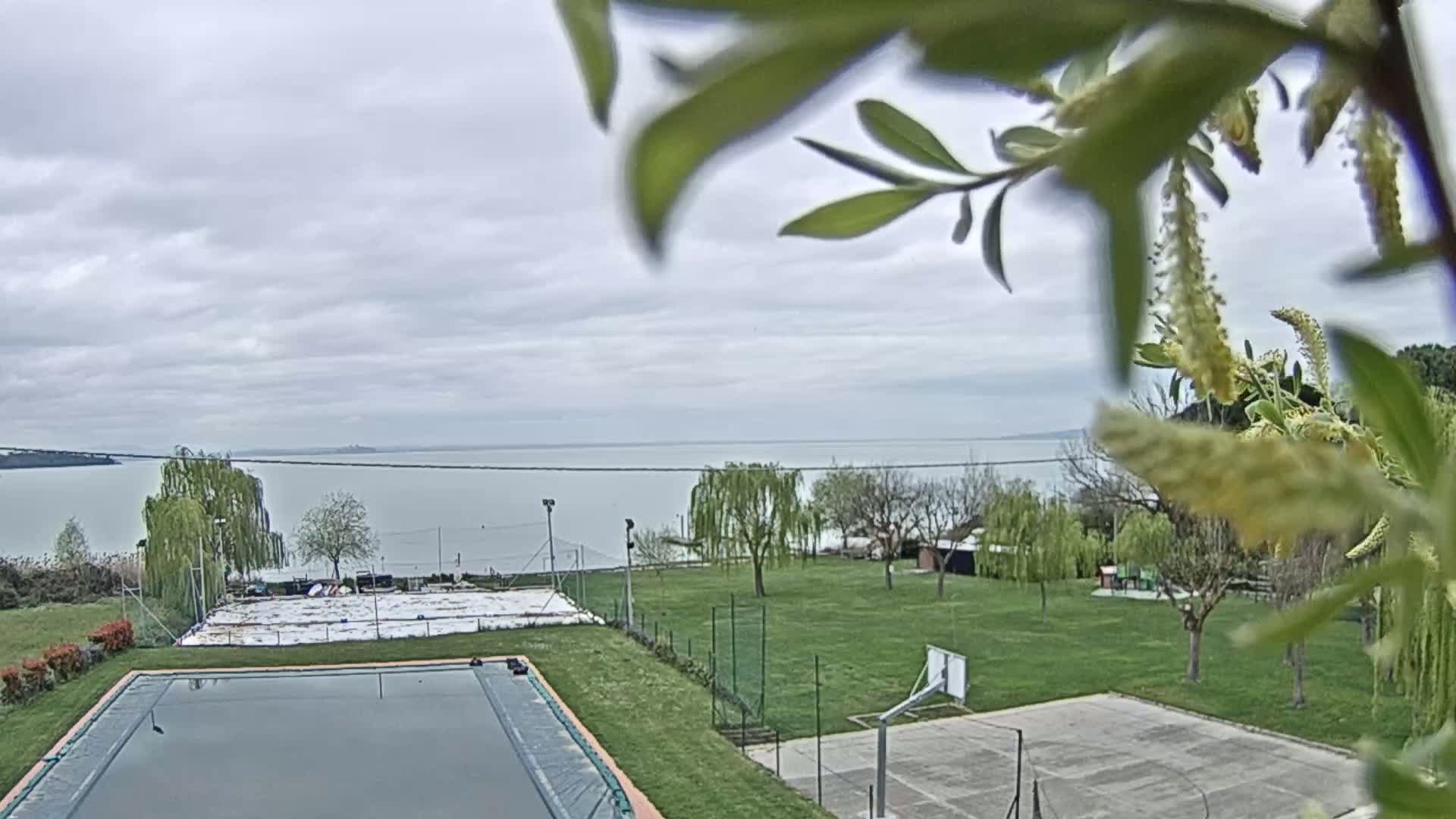 A mostly cloudy day overlooks a swimming pool, tennis court, basketball court, and grassy area with trees near a calm lake.