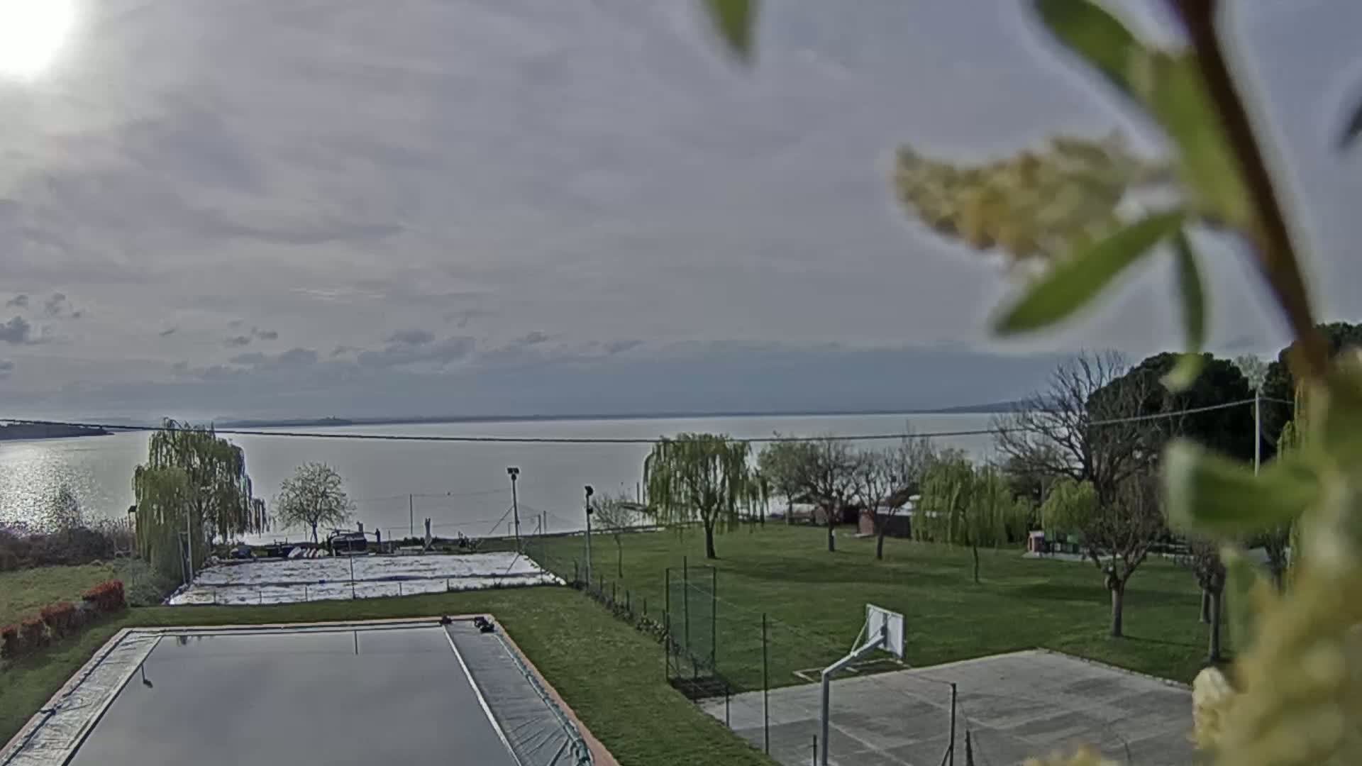A calm lake stretches across the horizon under a cloudy sky, viewed from a grassy area with a swimming pool, basketball court, and several trees.