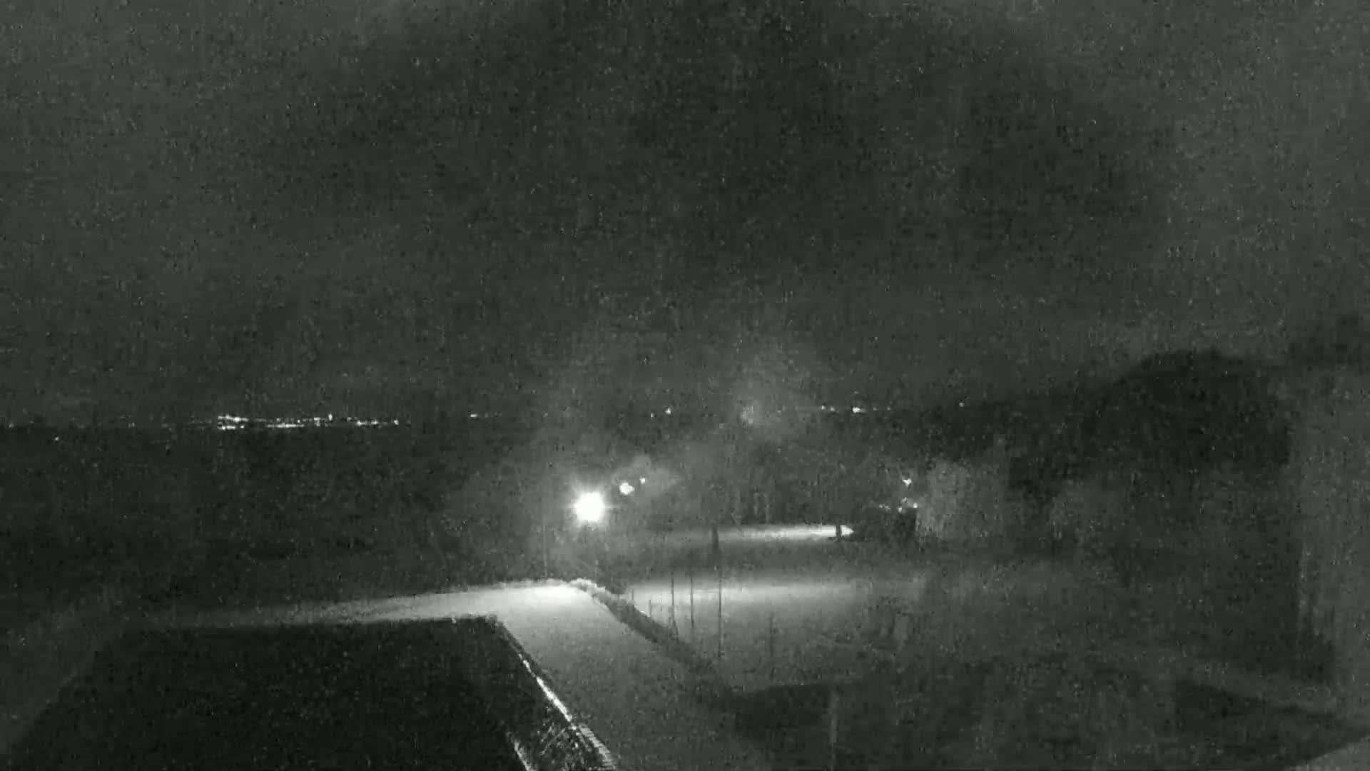 A nighttime, low-light view shows a snow-covered area with some structures and a distant, faintly lit horizon.