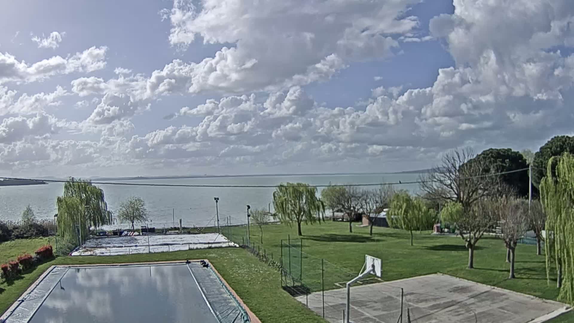 A partly cloudy sky overlooks a grassy area with a swimming pool, a basketball court, and a calm lake in the background.