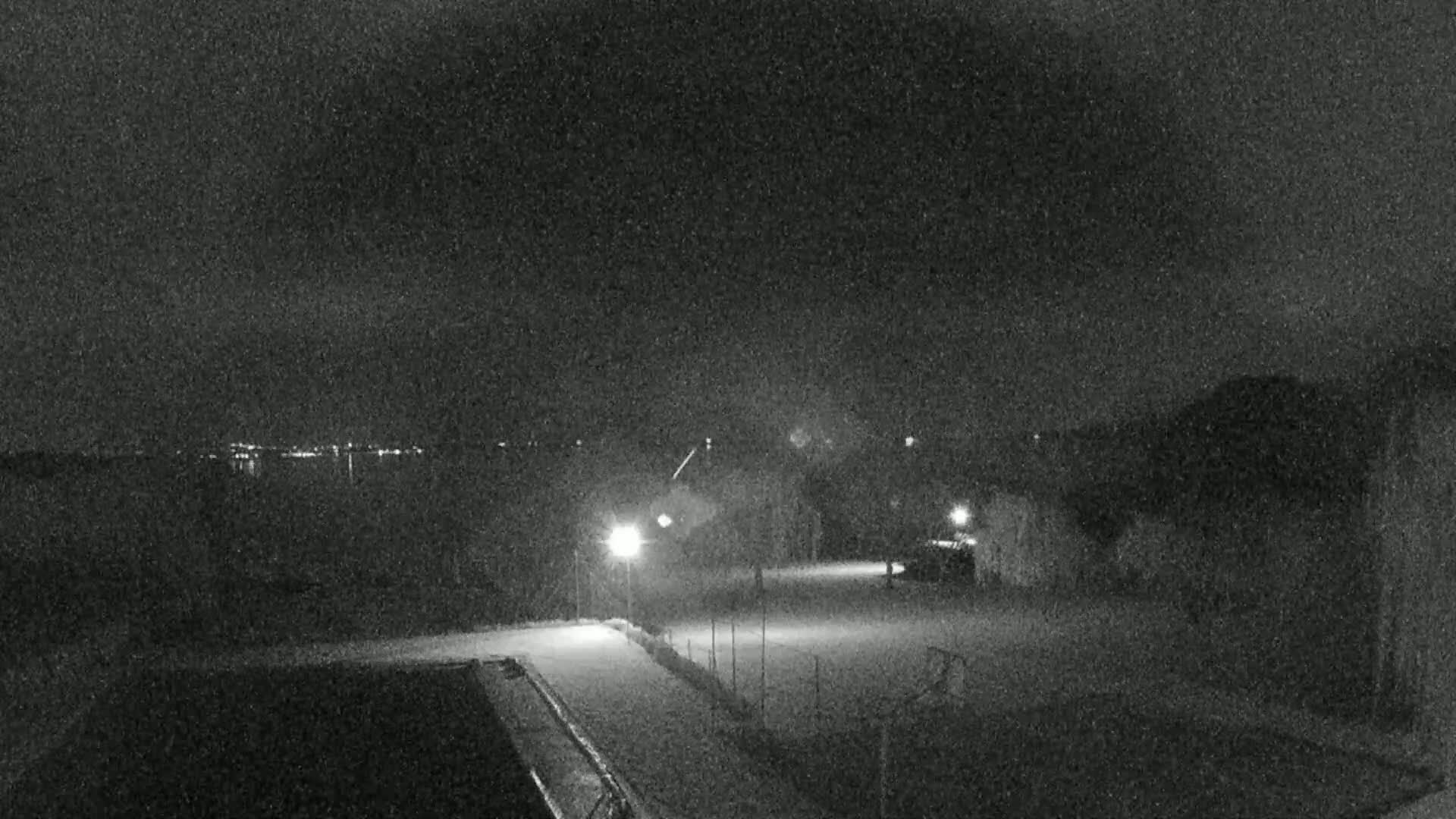 A nighttime, snow-covered outdoor scene shows a sparsely lit area with a building, distant city lights, and what appears to be a sports field.