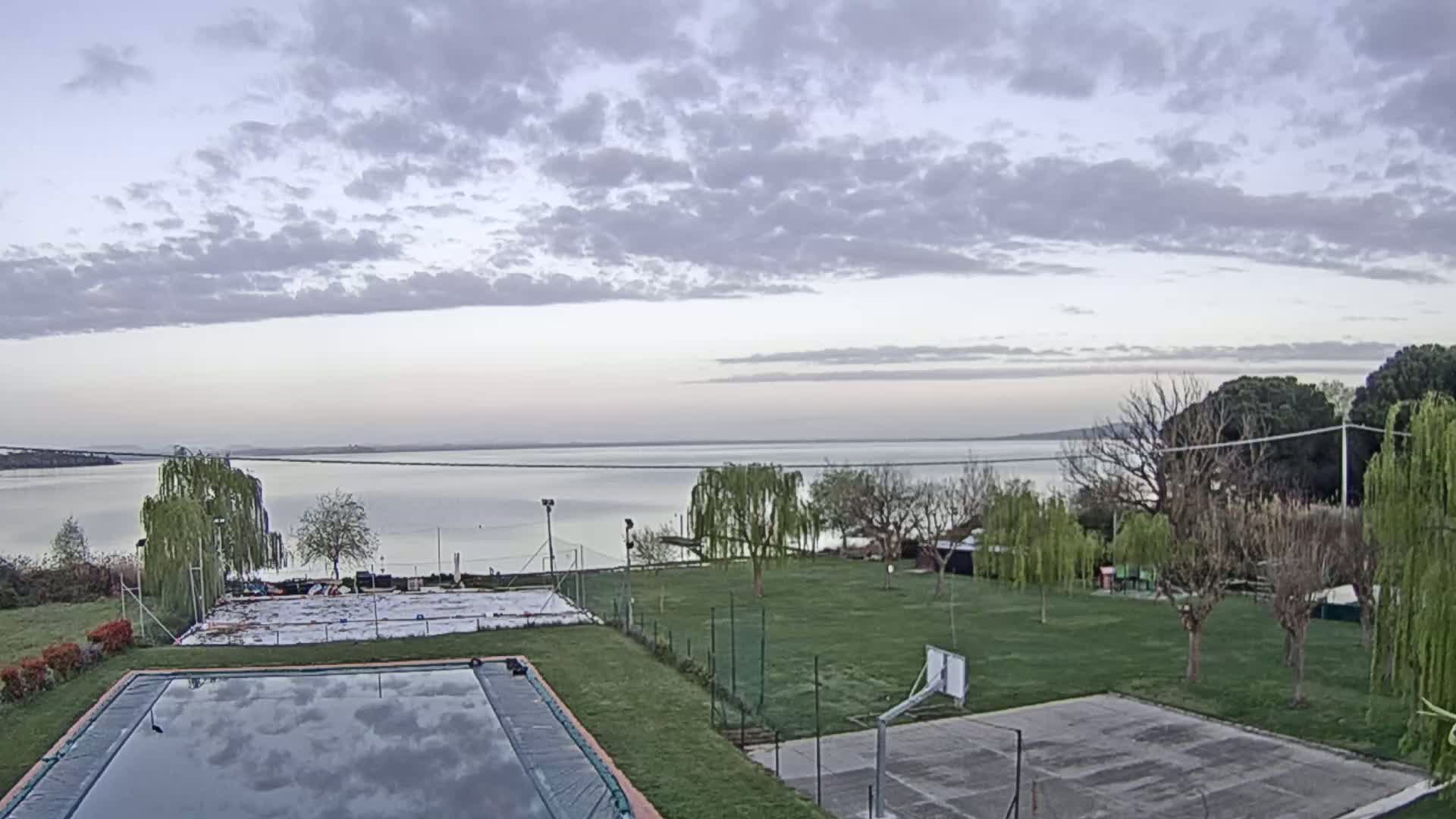 A mostly cloudy, calm day overlooks a large body of water, with a swimming pool and a basketball court situated on a grassy area in the foreground.