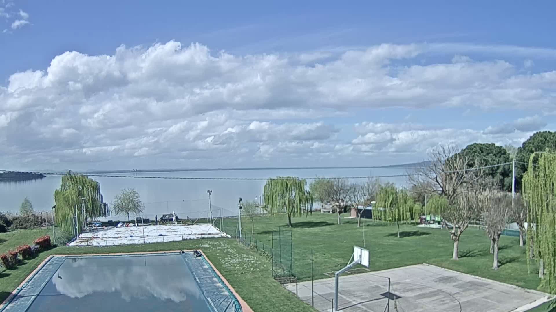 A swimming pool and basketball court sit on a grassy area overlooking a calm lake under a partly cloudy sky.