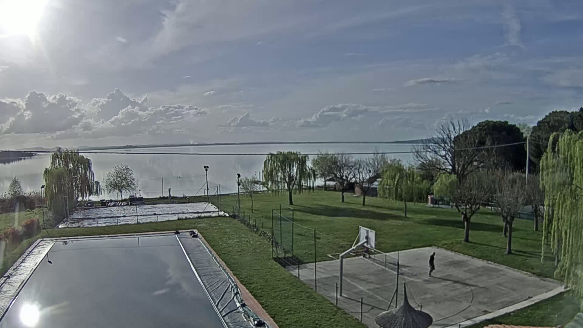A partly cloudy day overlooking a calm lake, with a swimming pool, basketball court, and grassy area in the foreground.