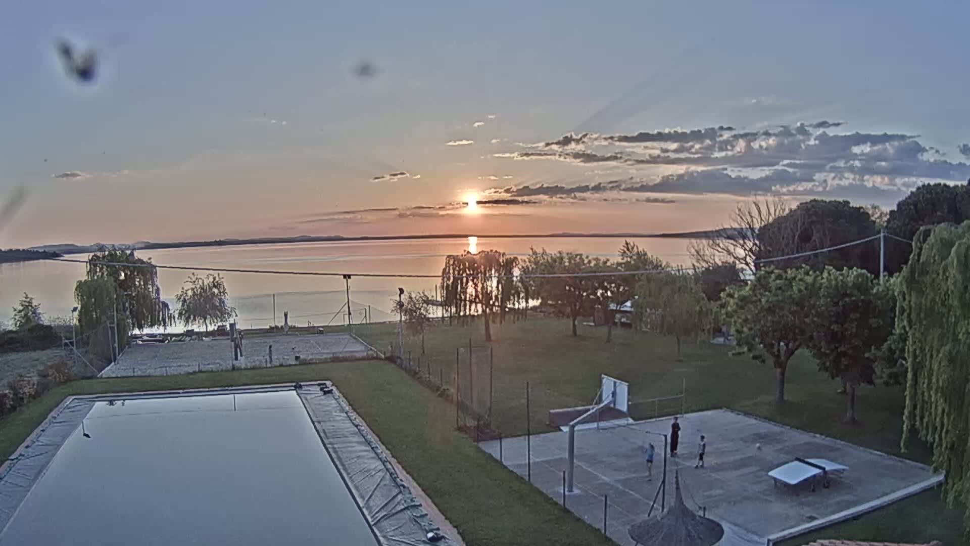 A tranquil sunset over a calm lake is viewed from above, showing a pool, basketball court, and volleyball court below, with a few people playing on the court.