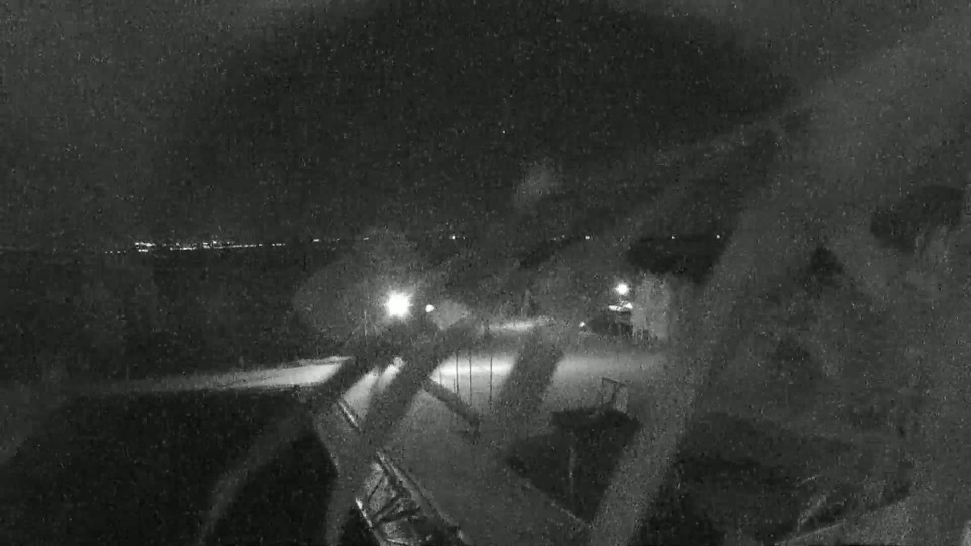 A nighttime, snowy scene shows a parking lot with some structures and distant lights.
