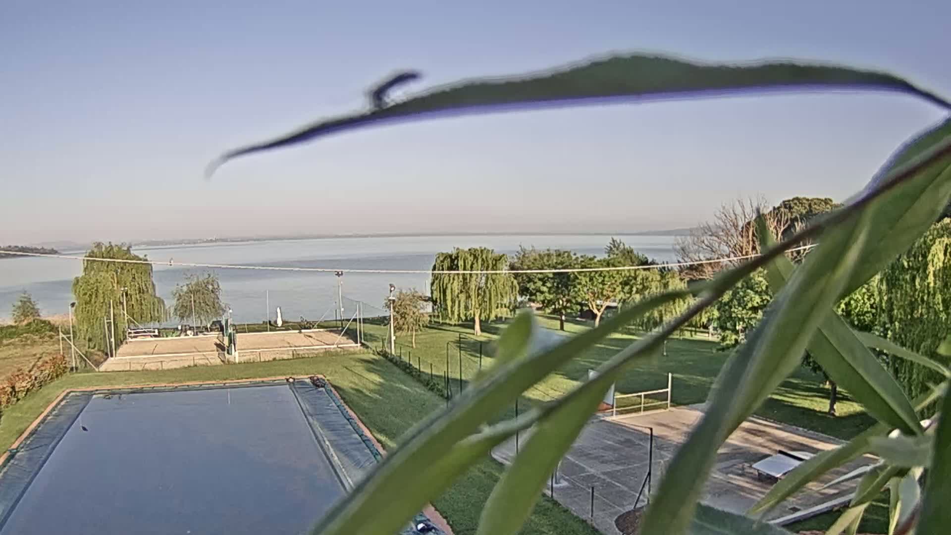 A swimming pool and tennis/basketball courts are situated on a grassy area overlooking a calm lake under a clear sky.