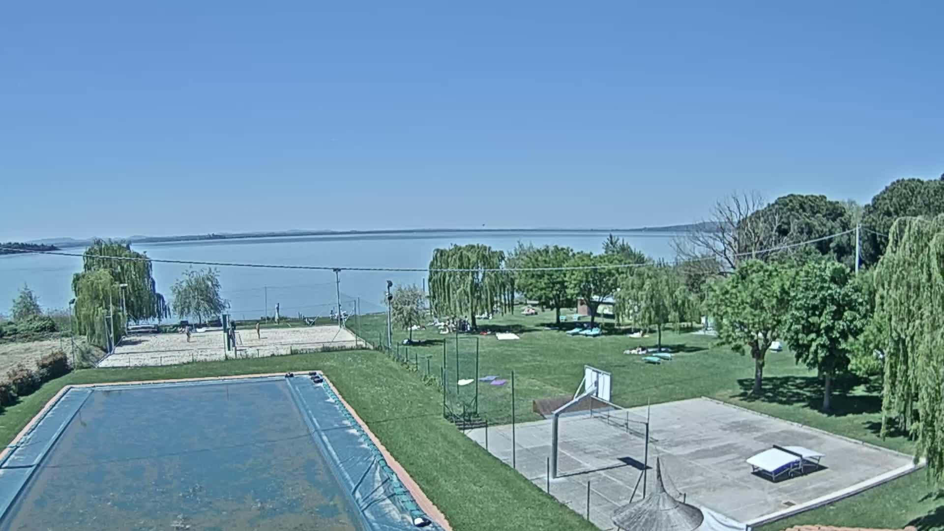 A sunny day overlooks a tranquil lake, showing a grassy area with a swimming pool, volleyball court, and basketball court.