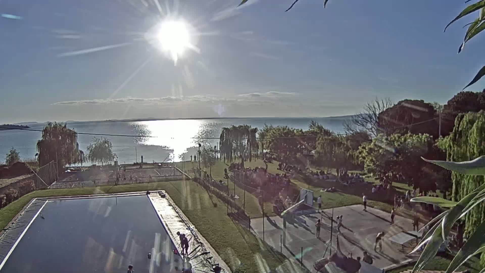 A sunny day overlooking a calm lake, with a grassy area containing a basketball court, a covered pool, and many people relaxing and playing sports.