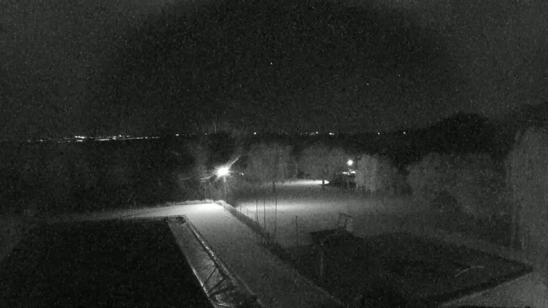 A nighttime, snow-covered sports field is illuminated by a few lights, with a distant cityscape faintly visible in the background.
