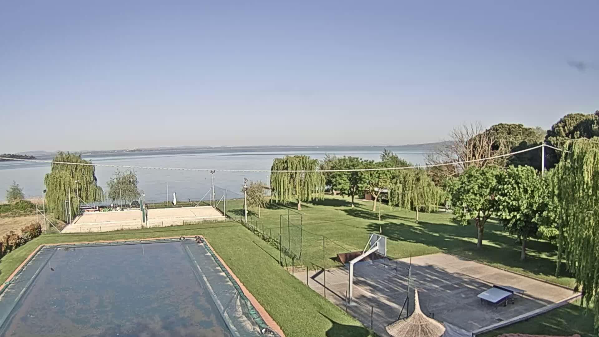 A calm lake is visible in the distance beyond a grassy area with a swimming pool, volleyball court, and basketball court under a clear, sunny sky.